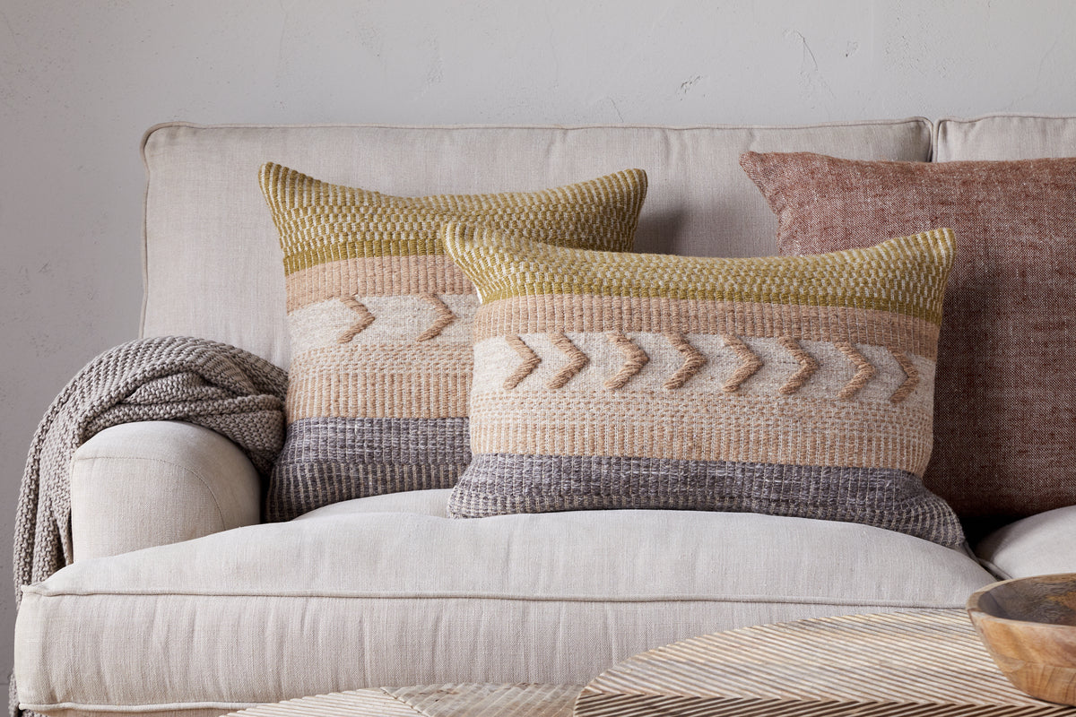 Two nkuku Tuleeka Wool Cushion Covers in ochre add texture to a light beige sofa, paired with a textured brown cover and a knitted throw. A wooden table sits in the foreground.