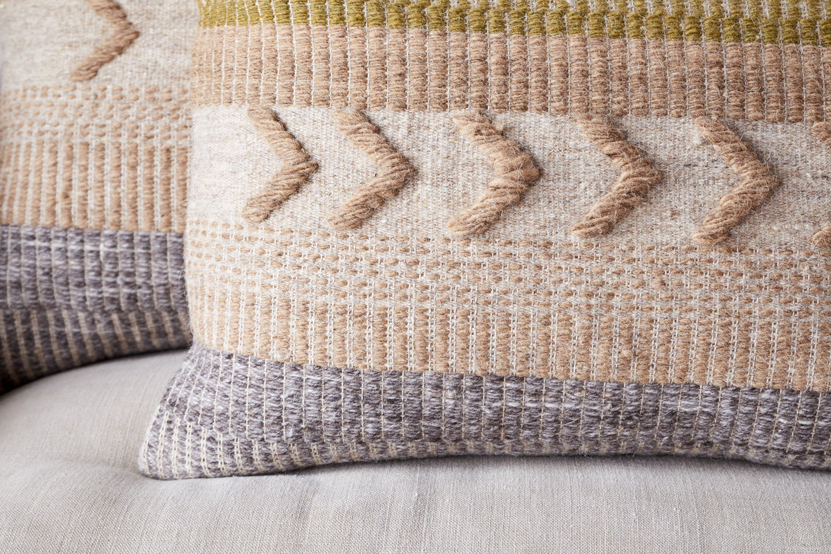 The Tuleeka Wool Cushion Cover - Ochre by nkuku features a textured woven design with geometric patterns in beige, gray, ochre, and olive green on a light background. Raised arrow shapes and stripes add depth to this decorative pillow.