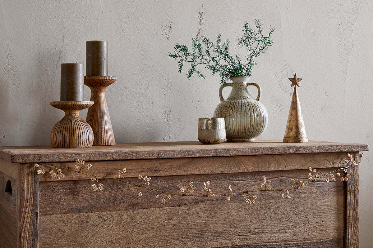 A wooden chest displays two tall ribbed candleholders with olive candles, a small ribbed vase with greenery, an antique brass votive, a gold cone-shaped tree topped with a star, and nkuku's Tanu Mango Wood Beaded Garland in Brass & Natural draped across the front.