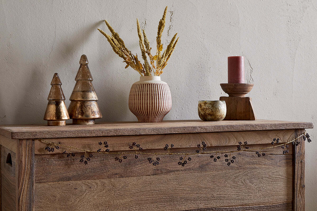 A rustic wooden chest is decorated with nkuku's Tanu Mango Wood Beaded Garland in Brass & Ruby, a ribbed beige vase with dried yellow grasses, gold ornaments, a red candle on wood, and a stone bowl against a textured beige wall.
