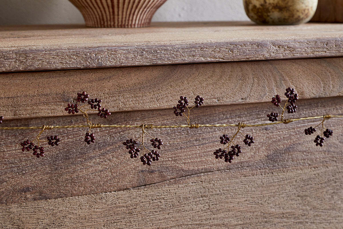 A close-up of the nkuku Tanu Mango Wood Beaded Garland in Brass & Ruby, featuring gold garland with berry-like beads on a rustic wooden surface, with blurred pottery vases in the background.