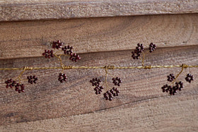 The nkuku Tanu Mango Wood Beaded Garland in Brass & Ruby features twisted gold wire and dark red beads, resembling festive berries, shown close-up against a textured wooden background.