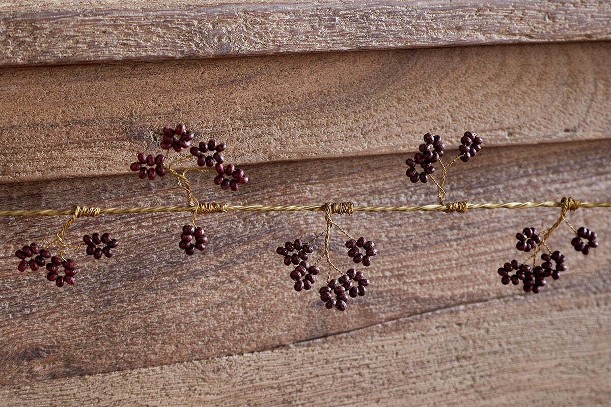 The nkuku Tanu Mango Wood Beaded Garland in Brass & Ruby features twisted gold wire and dark red beads, resembling festive berries, shown close-up against a textured wooden background.