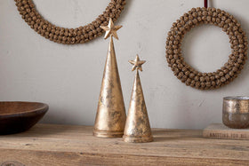 Two nkuku Talna Tree Bells in antique brass, shaped like metallic gold cones with star toppers, are displayed on a wooden surface among festive ornaments. In the background, two gold-decorated wreaths hang on a light wall.