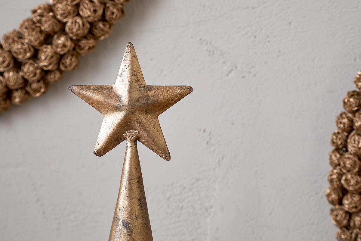 A close-up of the nkuku Talna Tree Bell in antique brass showcases its festive charm on a cone-shaped base. The background has a plain, light wall with decorative, braided wreaths partially visible on each side.