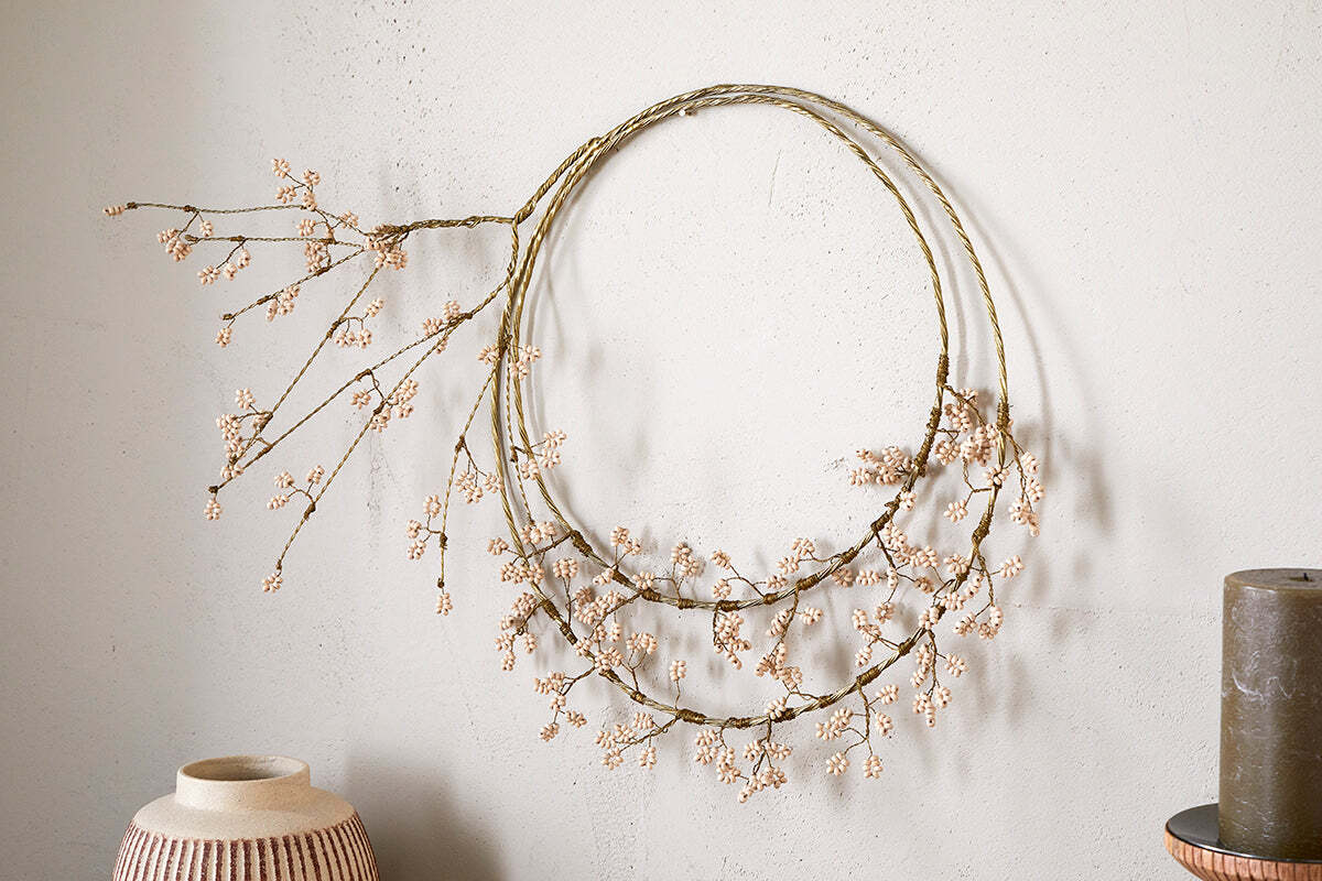 The Tanu Layered Mango Wood Beaded Wreath by nkuku, featuring brass and natural wood beads, hangs on a light textured wall. A ceramic vase and a dark cylindrical object are placed nearby for a stylish minimal look.