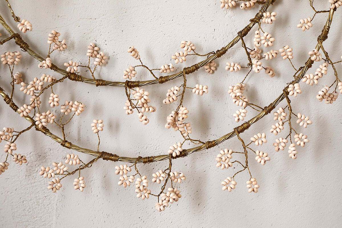 A close-up of the nkuku Tanu Layered Mango Wood Beaded Wreath in brass and natural, featuring overlapping loops of wood beads. Perfect for festive décor, it's displayed against a textured, off-white wall.