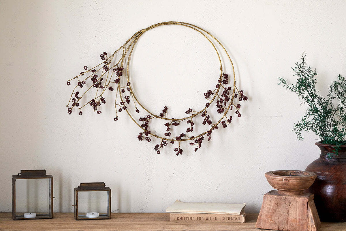 The nkuku Tanu Layered Mango Wood Beaded Wreath in Brass & Ruby hangs on a cream wall. Below, square lanterns, a brown vase with a plant, a stacked wooden bowl, and a book form an elegant festive centrepiece.