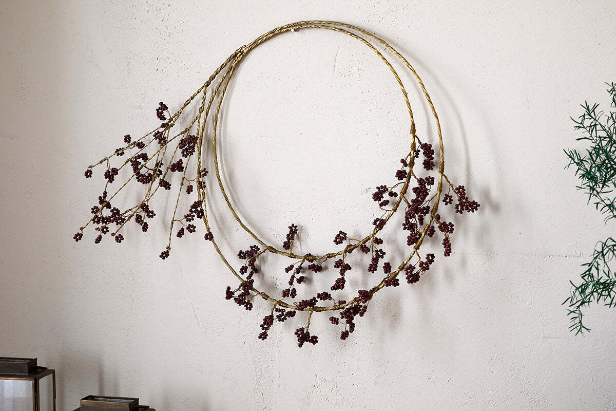 The Tanu Layered Mango Wood Beaded Wreath by nkuku features two slim brass rings with ruby red beads, hanging on a light textured wall. Green foliage appears on the right side of the image.