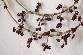 Close-up of two nkuku Tanu Layered Mango Wood Beaded Wreaths in Brass & Ruby, featuring twisted, branch-like designs and clusters of rich ruby beads for a rustic, antique-inspired look against a white textured wall.