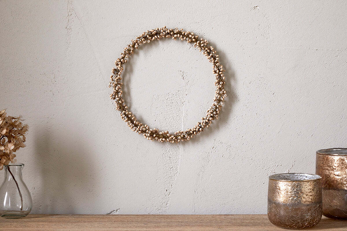 The nkuku Tanu Mango Wood Beaded Wreath in Brass & Natural hangs on a light, textured wall, paired with metallic candle holders and a clear vase of dried flowers for an elegant and minimalist Christmas display.