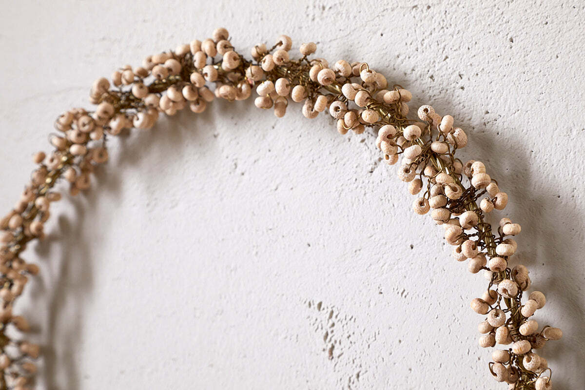 The nkuku Tanu Mango Wood Beaded Wreath in brass and natural features minimalist wooden beads for a rustic Christmas touch, shown hanging on a textured off-white wall.