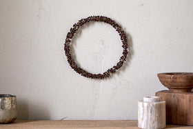 A minimalist wall features the nkuku Tanu Mango Wood Beaded Wreath in Brass & Ruby. Below, a wooden surface displays a silver cup, marble container, antique brass bowl, and wooden box for an earthy look.