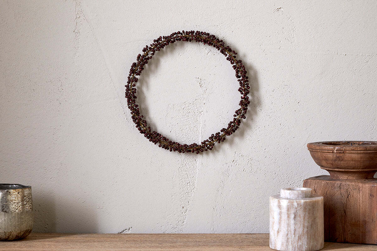 A minimalist wall features the nkuku Tanu Mango Wood Beaded Wreath in Brass & Ruby. Below, a wooden surface displays a silver cup, marble container, antique brass bowl, and wooden box for an earthy look.