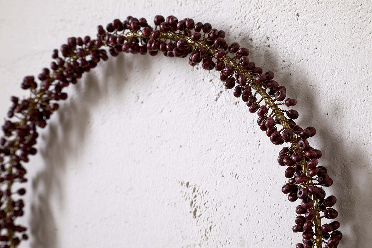 A partial view of nkuku's Tanu Mango Wood Beaded Wreath in Brass & Ruby, featuring wood beads and deep red accents, hangs on a textured white wall—ideal for Christmas decor.