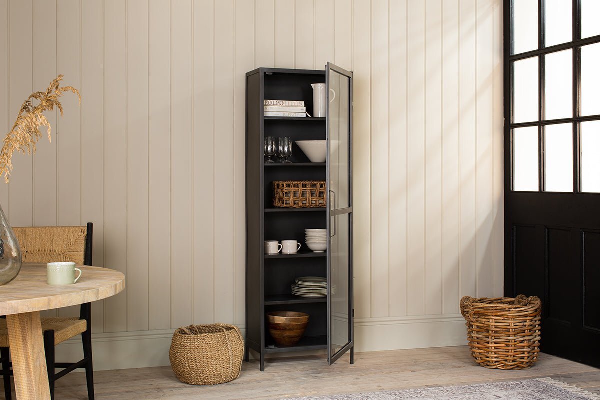 The Takua Iron Cabinet - Narrow by nkuku stands on light wood flooring, its glass door revealing shelves with dishes, bowls, cups, and baskets. Nearby are woven baskets, a round table with a chair, and a windowed black door against cream paneled walls.