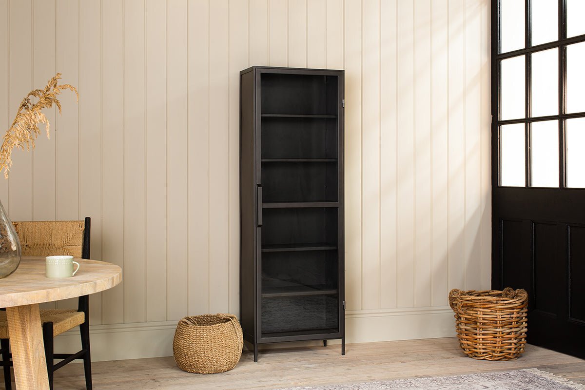The Takua Iron Cabinet - Narrow by nkuku stands against a beige paneled wall. Nearby, a round wooden table with a mug, woven chair, and two wicker baskets rest on a light wood floor by a large window and black door. Dried grass decorates the table.