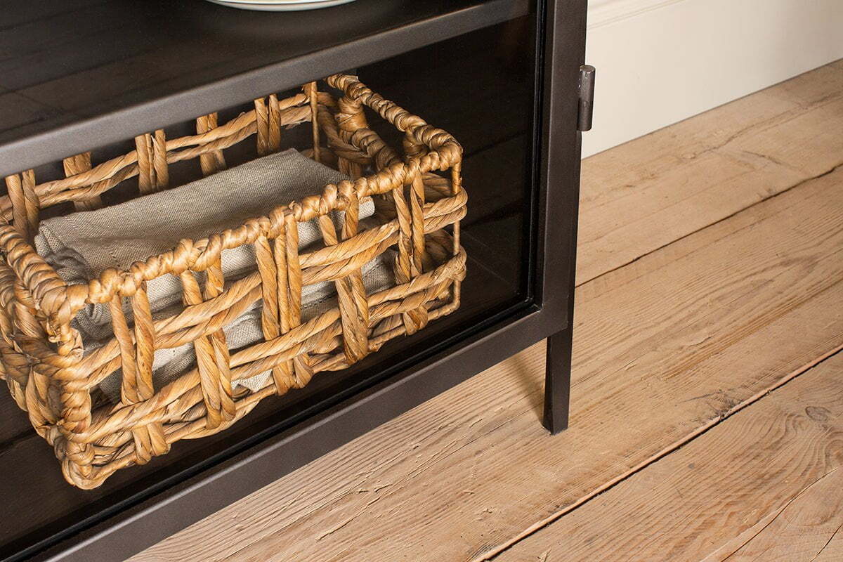 A woven wicker basket with tan fabric rests on a shelf of the nkuku Takua Iron Cabinet – Narrow, placed on a rustic wooden floor.