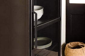 Close-up of the Takua Iron Cabinet - Narrow by nkuku, featuring a black glass front and metal handle. White bowls are on the top shelf and plates on the lower, with a woven basket nearby beside a black door with a white window.