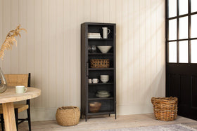 The nkuku Takua Iron Cabinet - Narrow, with black framing and glass doors, stands against a cream-paneled wall holding dishes, bowls, baskets, and a pitcher. Nearby are woven baskets, a round table with a chair, and a large windowed black door.