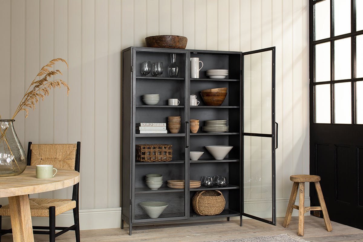 The nkuku Takua Iron Cabinet - Wide, finished in black with glass doors, showcases dishes, bowls, glasses, and woven baskets. It stands against a cream paneled wall in a sunlit room featuring a wooden table and woven chair near a windowed black door.
