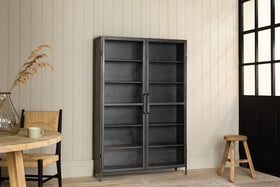 The Takua Iron Cabinet - Wide by nkuku, featuring glass fronts and five shelves, stands against cream panel walls. A light wood table, chair, glass vase with dried grasses, wooden stool, and black-paned door complete the minimalist space.