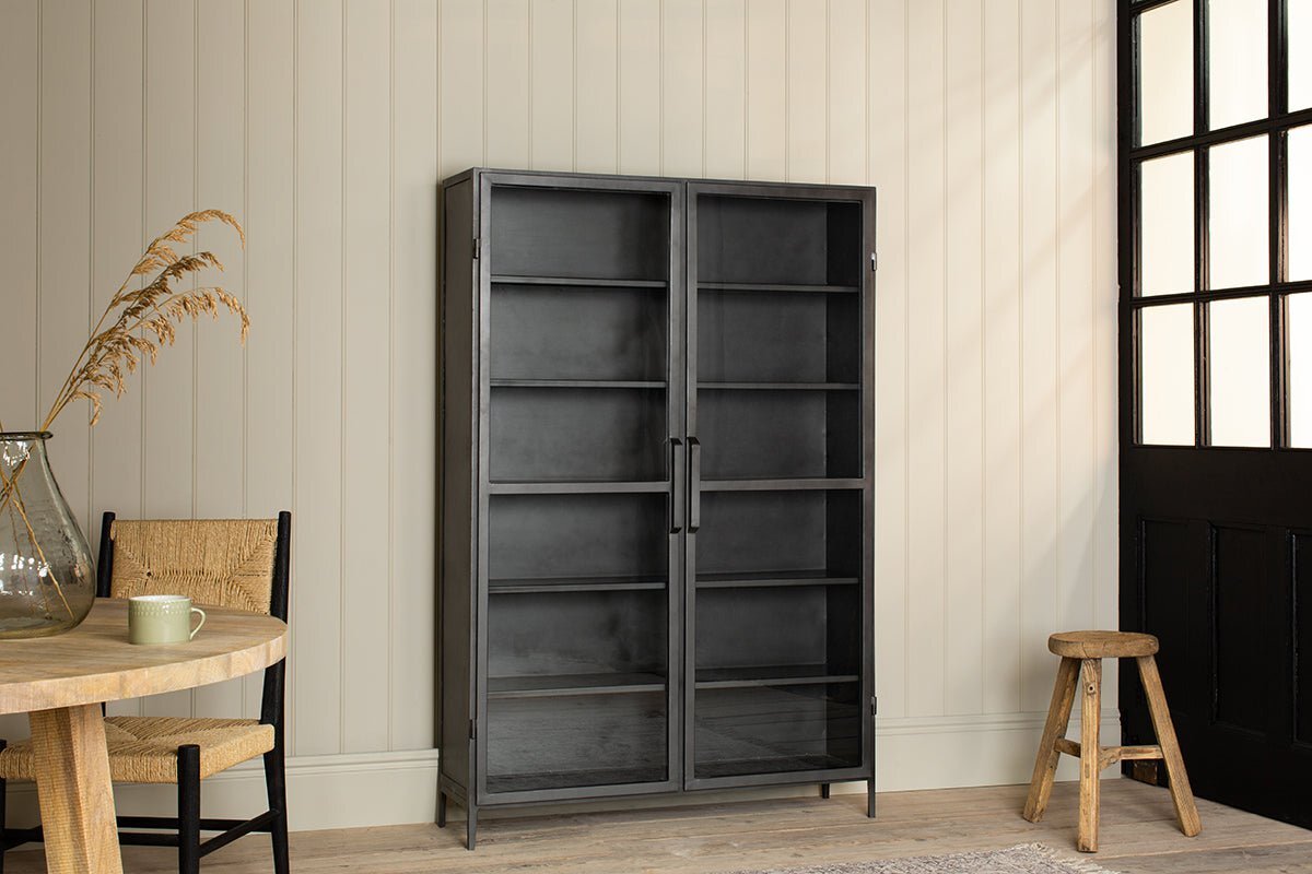 The Takua Iron Cabinet - Wide by nkuku, featuring glass fronts and five shelves, stands against cream panel walls. A light wood table, chair, glass vase with dried grasses, wooden stool, and black-paned door complete the minimalist space.