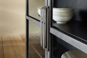 A close-up of the nkuku Takua Iron Cabinet - Wide, featuring a modern handle and glass front, displays stacks of white textured bowls on dark shelves. Light walls and wooden floorboards are seen in the background.