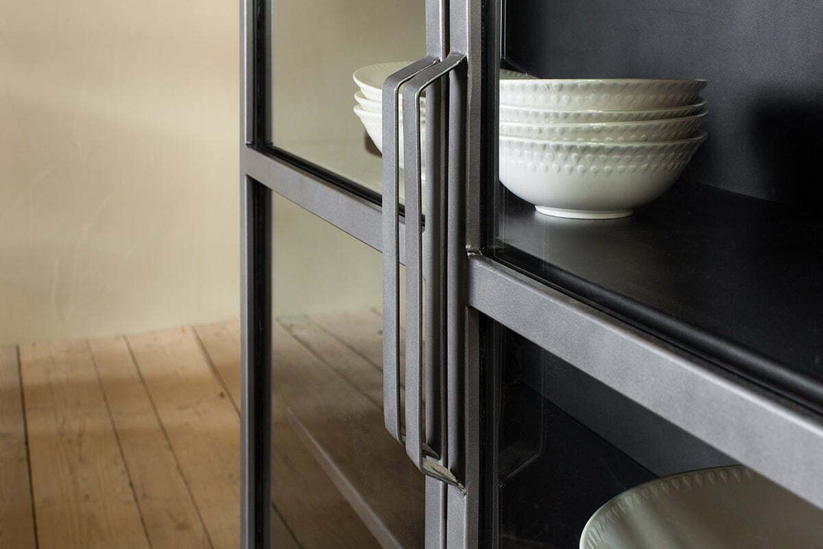 A close-up of the nkuku Takua Iron Cabinet - Wide, featuring a modern handle and glass front, displays stacks of white textured bowls on dark shelves. Light walls and wooden floorboards are seen in the background.