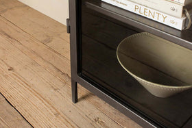 The Takua Iron Cabinet - Wide by nkuku showcases a beige bowl on a dark wooden shelf behind its glass door, with books arranged above—offering a minimalist blend of style and function.