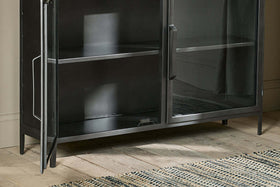 A close-up of the nkuku Takua Iron Sideboard, an industrial black metal and glass cabinet with two glass doors—one ajar—showing two shelves inside. The sideboard stands on four legs on a light wooden floor beside a cream and dark blue woven rug.