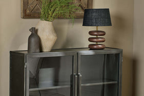 The Takua Iron Sideboard by nkuku stands against a beige wall, topped with a tall beige textured vase of green foliage, a smaller brown bottle-shaped vase, and a table lamp featuring a black shade and sculptural wooden base.