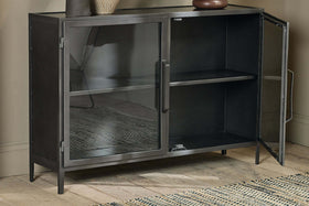 The Takua Iron Sideboard by nkuku, featuring an industrial black metal design and two glass doors, stands on wooden flooring against a beige wall. It has a single interior shelf, with a vase and bowl on top and a woven rug partially visible on the left.