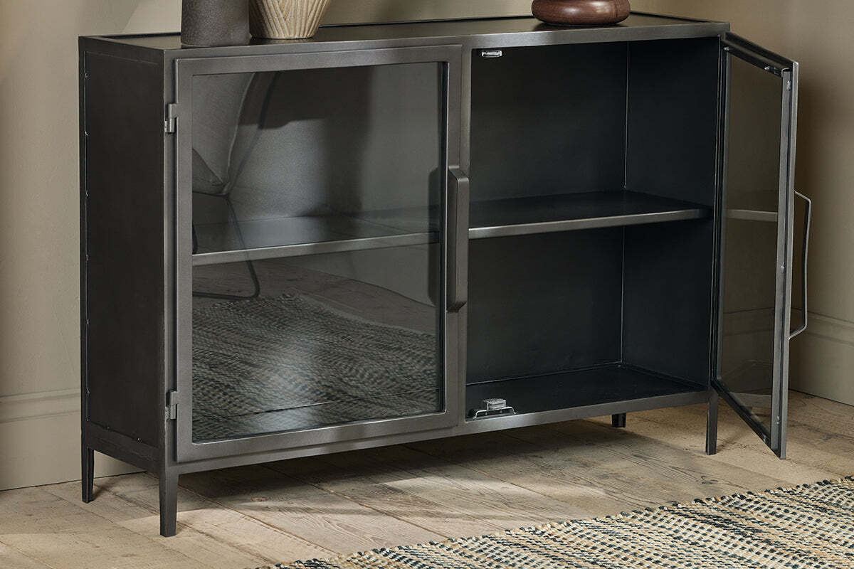 The Takua Iron Sideboard by nkuku, featuring an industrial black metal design and two glass doors, stands on wooden flooring against a beige wall. It has a single interior shelf, with a vase and bowl on top and a woven rug partially visible on the left.