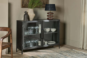 The nkuku Takua Iron Sideboard displays glassware and white dishes behind a glass front. On top are two vases—one ribbed with greenery, one dark and narrow—and a black-shaded modern lamp, alongside a wood chair and patterned rug.