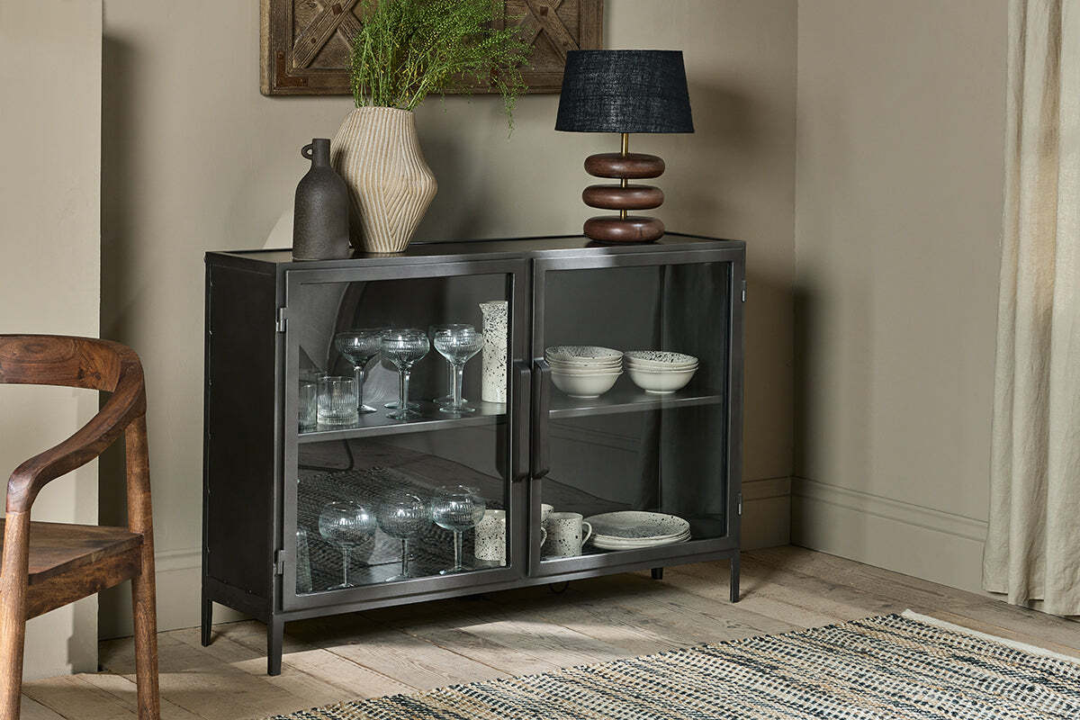 The nkuku Takua Iron Sideboard displays glassware and white dishes behind a glass front. On top are two vases—one ribbed with greenery, one dark and narrow—and a black-shaded modern lamp, alongside a wood chair and patterned rug.