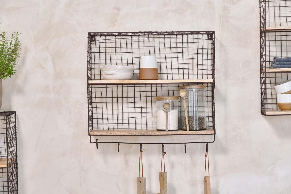 The nkuku Tamba Shelf with Hooks, finished in rust, is wall-mounted and displays stacked bowls, a cup, jars with lids, and salt and pepper shakers. Utensils hang from the hooks below against a neutral textured wall with plants and another shelf nearby.
