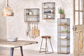 A cozy kitchen features two nkuku Tamba Wall Hung Shelves on a textured beige wall—one with hanging hooks for towels and boards. The shelves display dishes, bowls, and a potted plant above a wooden table and stool.