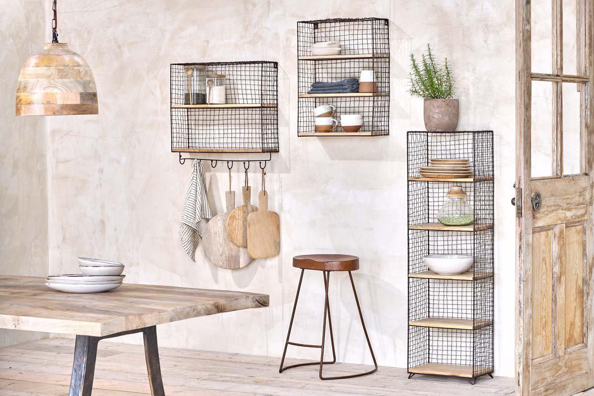 A cozy kitchen features two nkuku Tamba Wall Hung Shelves on a textured beige wall—one with hanging hooks for towels and boards. The shelves display dishes, bowls, and a potted plant above a wooden table and stool.