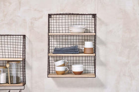 The nkuku Tamba Wall Hung Shelf, with three wire shelves, displays stacked ceramic mugs, a bowl, a dish, and two folded blue cloths on a light beige wall. Another shelf with jars and utensils is partially visible to the left.