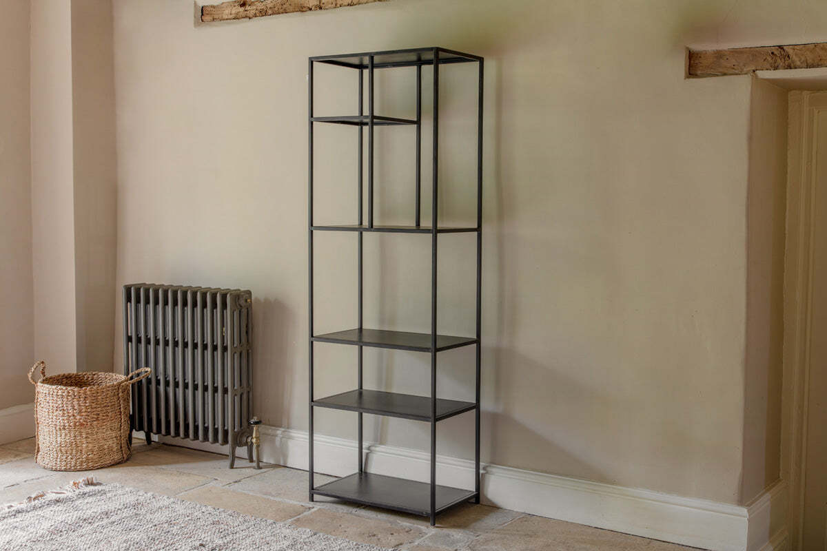 The Tanvi Display Shelf - Black - Narrow by nkuku stands against a beige wall. With five open shelves, it’s next to a gray radiator and woven basket, on tiled flooring with part of a rug beneath exposed wooden beams above.