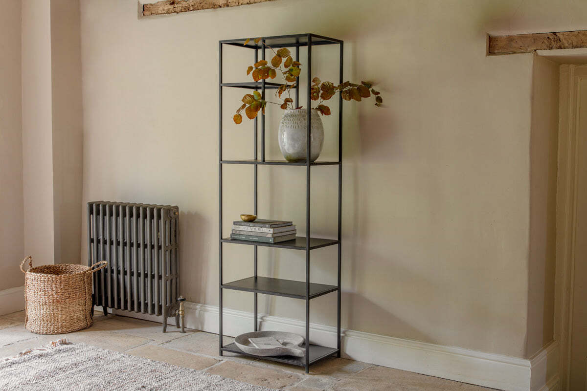 The nkuku Tanvi Display Shelf - Black - Narrow, with five shelves and a sleek metal frame, stands against a cream wall holding autumn leaves, books, and decor. Beside it are a vintage grey radiator and woven basket on the stone floor.