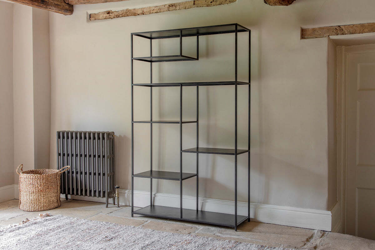 The Tanvi Display Shelf - Black - Wide by nkuku stands against a beige wall near a gray radiator and woven basket. Wooden ceiling beams, a door, and a light rug on stone flooring add to the room's simple, rustic charm.