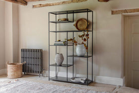 The Tanvi Display Shelf - Black - Wide by nkuku showcases vases, books, and a wooden tray. To its left, a grey radiator and woven basket sit on the stone floor.