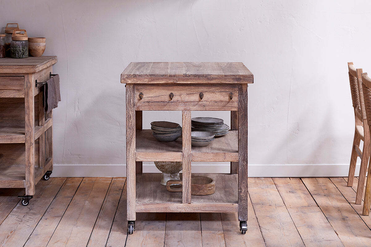 The Tivu Reclaimed Wood Butchers Block by nkuku features a thick top, open shelves, and wheeled legs. Set on a wooden floor before a white textured wall, this farmhouse cart adds rustic charm to any kitchen.
