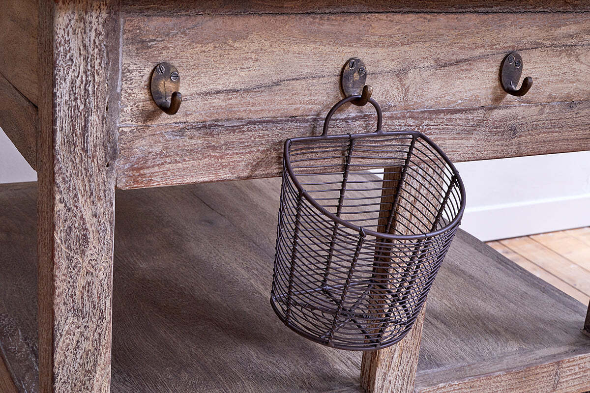 The nkuku Tivu Reclaimed Wood Butchers Block - Natural features a weathered finish, thick legs, lower shelf, and metal hooks beneath the tabletop. A small angled wire basket hangs from one hook, with side lighting highlighting the wood grain.