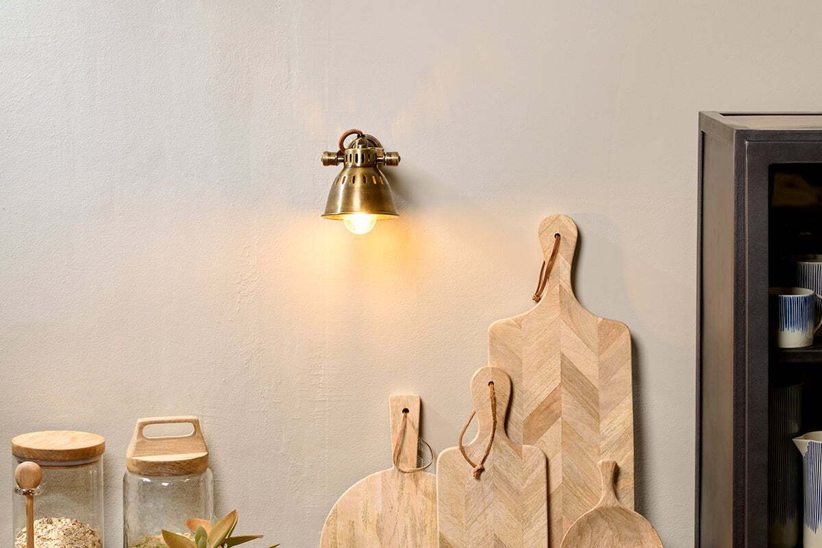 The nkuku Tubu Brass Spot Light in Antique Brass casts warm lighting on a beige wall above wooden cutting boards. Nearby are a glass jar with a wooden lid, a potted plant, and part of a dark cabinet.