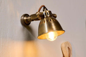The nkuku Tubu Brass Spot Light in Antique Brass, inspired by industrial design, features a glowing bulb and vented shade. Its brown fabric cord casts warm yellow light and soft shadows on beige walls for a timeless touch.
