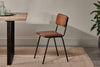 The Ukari Leather Dining Chair - Aged Tan by nkuku, featuring a padded leather seat and metal frame, sits beside a wooden table on a beige woven rug. Neutral tones and a visible plant add to the space's modern, utilitarian style.