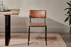 The Ukari Leather Dining Chair - Aged Tan by nkuku, featuring a padded seat, square backrest, and slim black metal legs, sits on a beige rug beside a wooden table with glassware and a potted plant against a light wall.